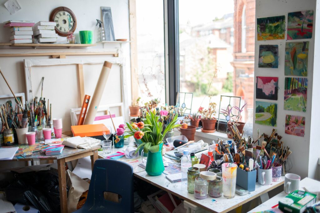 a bright studio featuring a desk in front of a window. the desk is full of flowers, pant and other creative tools.